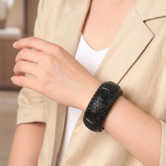 A close-up of a person wearing a thick, textured black bangle bracelet on their wrist, styled with a beige blazer and white top.