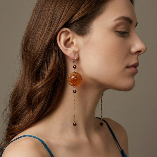 A woman in profile wearing a long amber-orange beaded drop earring with gold-toned chain details, against a soft taupe background.