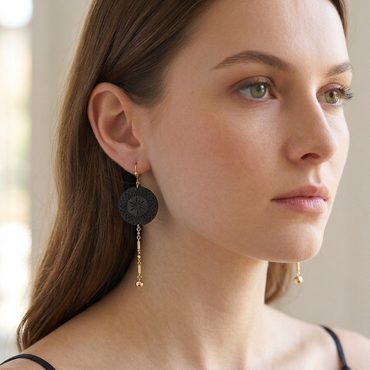 A woman wearing a pair of long black circular drop earrings with gold-toned chain details, shown in a close-up portrait against a soft, light background.