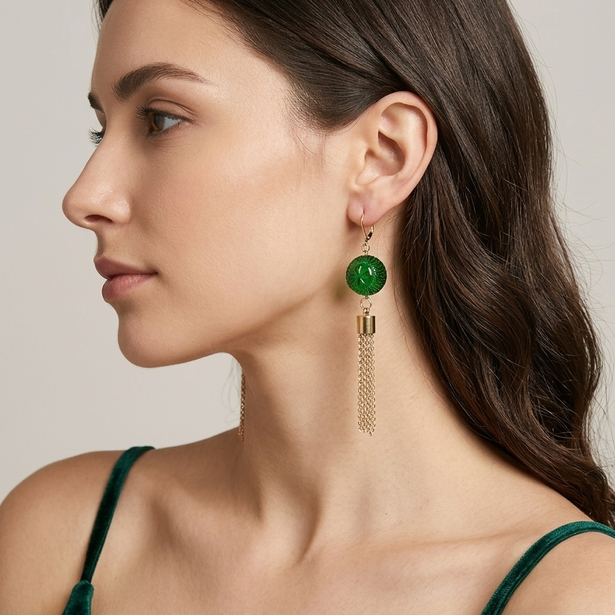 A woman in profile wearing a dangling earring with a round green stone and gold-tone chain tassels, paired with a dark green velvet top against a light beige background.