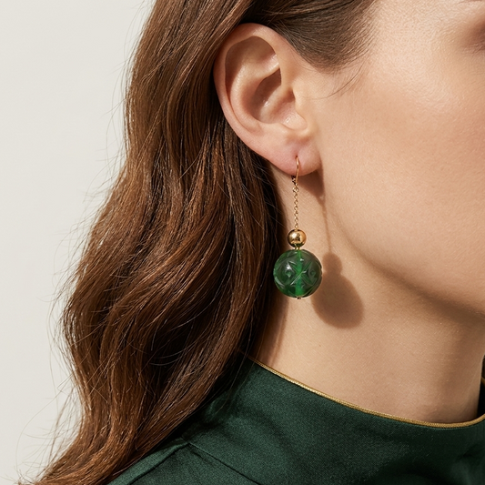 A close-up of a woman wearing a dangling earring with a round green stone and a small gold bead on a delicate gold chain, paired with a dark green high-neck top.