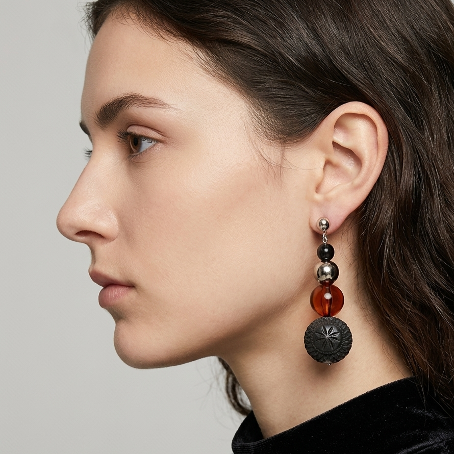 A close-up side profile of a woman wearing a long, multi-tiered dangle earring. The earring features a silver-toned post with black and metallic beads, a translucent reddish-brown bead, and a large textured dark gray disc at the bottom.