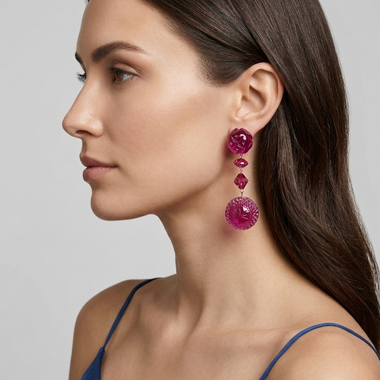 A close-up side profile of a woman with long dark brown hair wearing a pair of chunky, multi-tiered magenta pink dangle earrings. She is wearing a blue satin spaghetti strap top against a neutral light gray background.