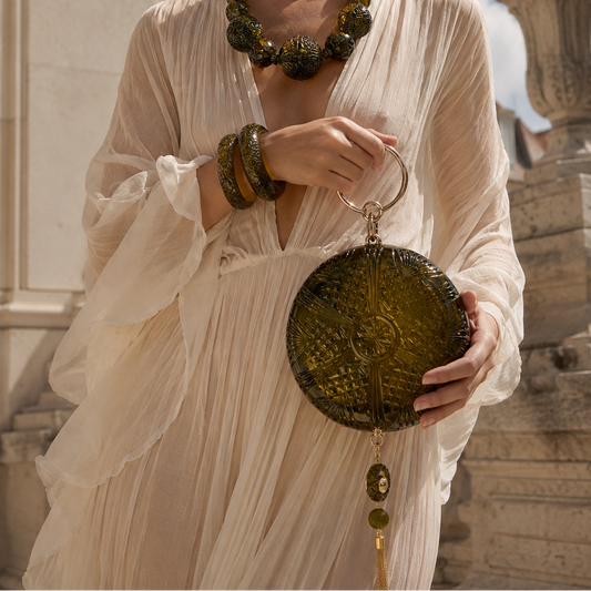 A woman in a cream-colored draped dress holds a round, textured olive green clutch bag with a tassel. She is accessorized with a matching olive green beaded necklace and bangles, set against a background of a stone building and blue sky.