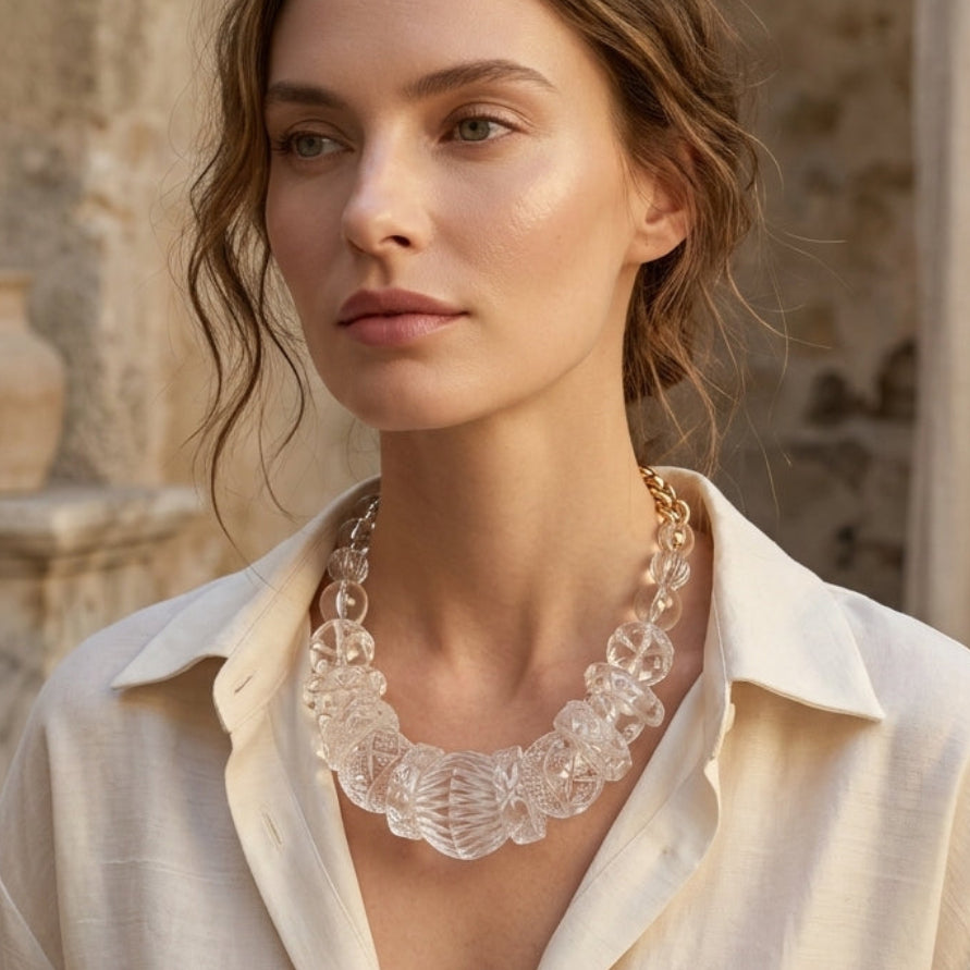 A woman wearing a chunky, clear resin statement necklace with a scalloped edge, styled with a cream linen shirt in an outdoor setting with stone columns.