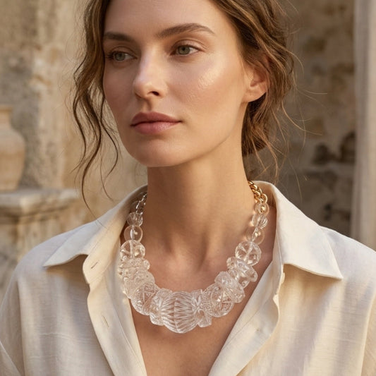 A woman wearing a chunky, clear resin statement necklace with a scalloped edge, styled with a cream linen shirt in an outdoor setting with stone columns.