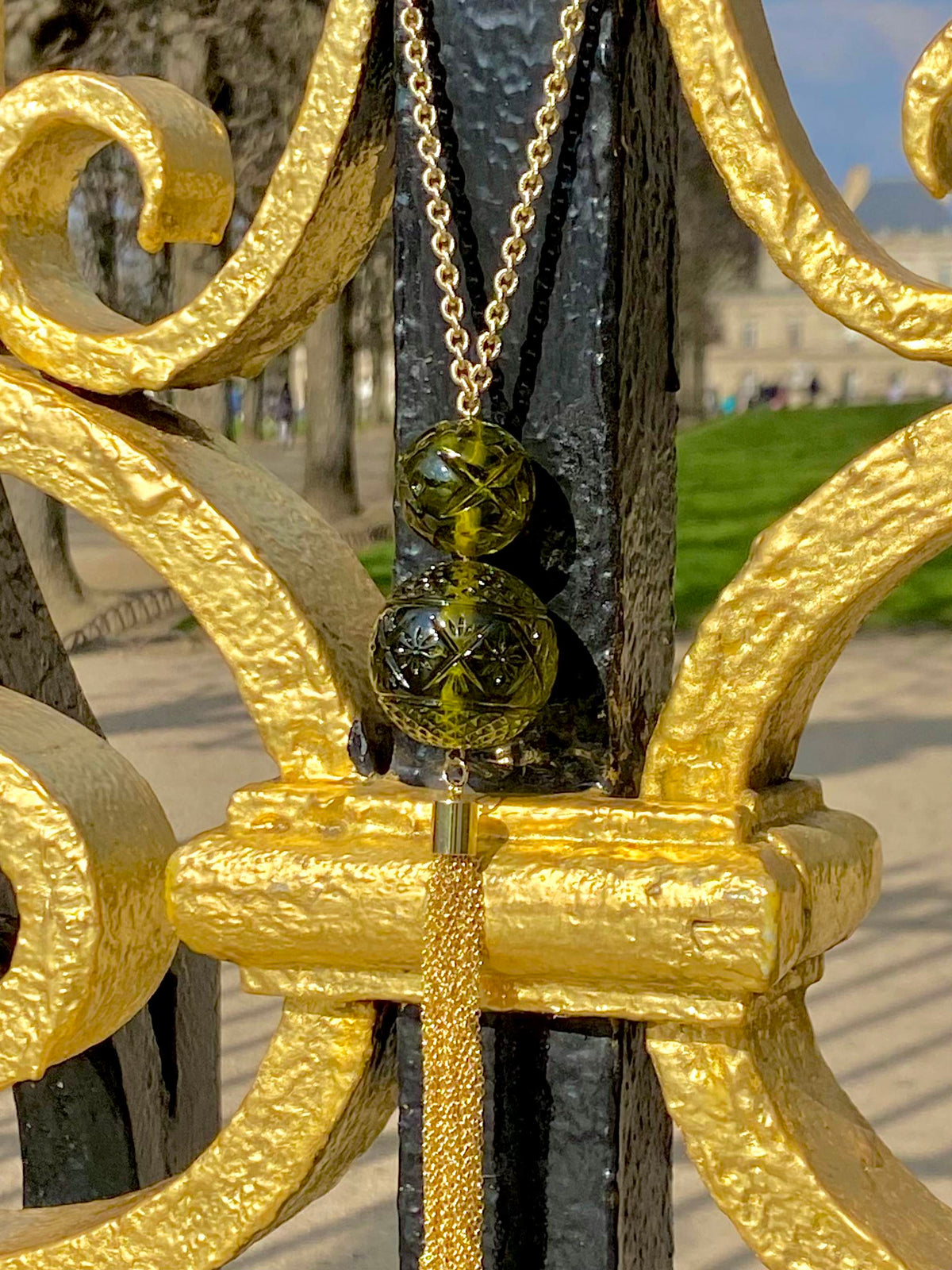 A gold chain necklace with two olive green glass beads and a gold tassel, hanging from an ornate gold and black metal fence in an outdoor park setting.