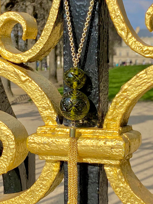 A gold chain necklace with two olive green glass beads and a gold tassel, hanging from an ornate gold and black metal fence in an outdoor park setting.