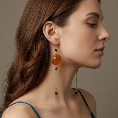 A woman in profile wearing a long amber-orange beaded drop earring with gold-toned chain details, against a soft taupe background.