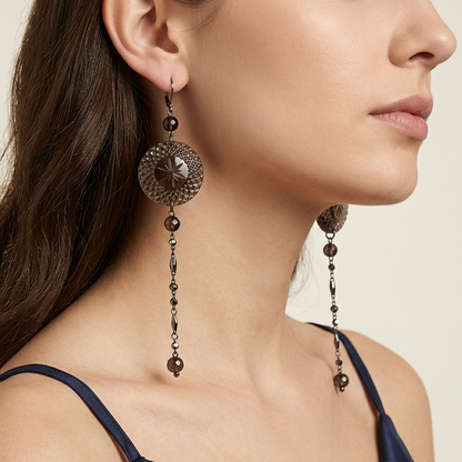 A close-up side profile of a woman wearing ornate dangling earrings with circular filigree tops and long beaded drops in a dark bronze finish, paired with a navy camisole against a light beige background.