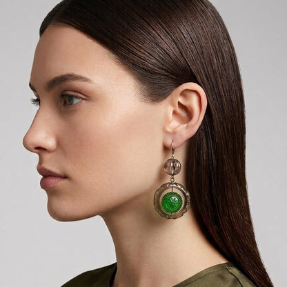 A woman in profile wearing a dangling earring with a round green stone and textured metallic frame, shown against a light gray studio background.