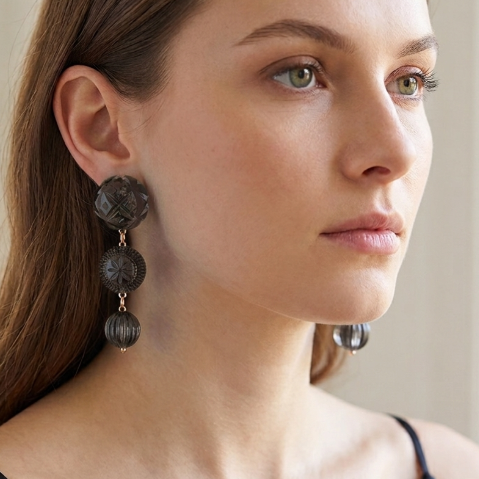 A close-up of a woman wearing dark charcoal textured drop earrings with circular disc charms and small gold-toned connectors, set against a soft, bright, out-of-focus indoor background.