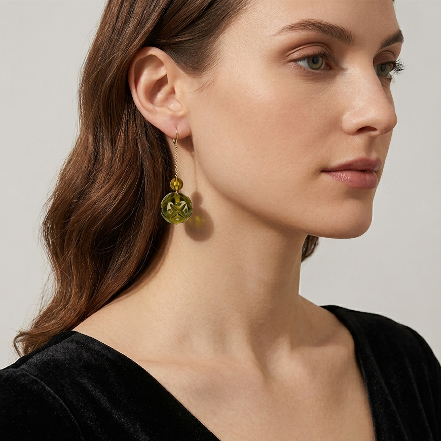 A close-up of a woman wearing a dangling earring with a faceted magenta-pink gemstone and a gold-toned post, paired with a black velvet top against a light gray background.