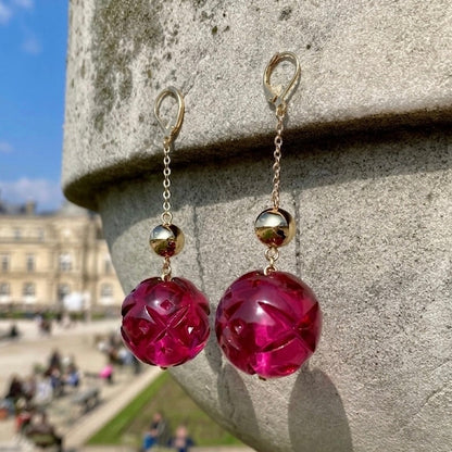 A pair of dangling earrings with faceted magenta-pink round beads and small metallic gold-toned spheres on chains, photographed hanging against a stone structure outdoors with a blurred city scene in the background.