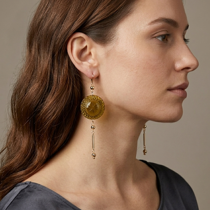 A woman in profile wearing large drop earrings with round amber-yellow textured discs and dangling gold-toned beads, against a soft beige background.