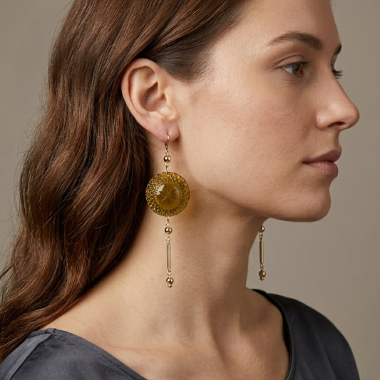 A woman in profile wearing large drop earrings with round amber-yellow textured discs and dangling gold-toned beads, against a soft beige background.