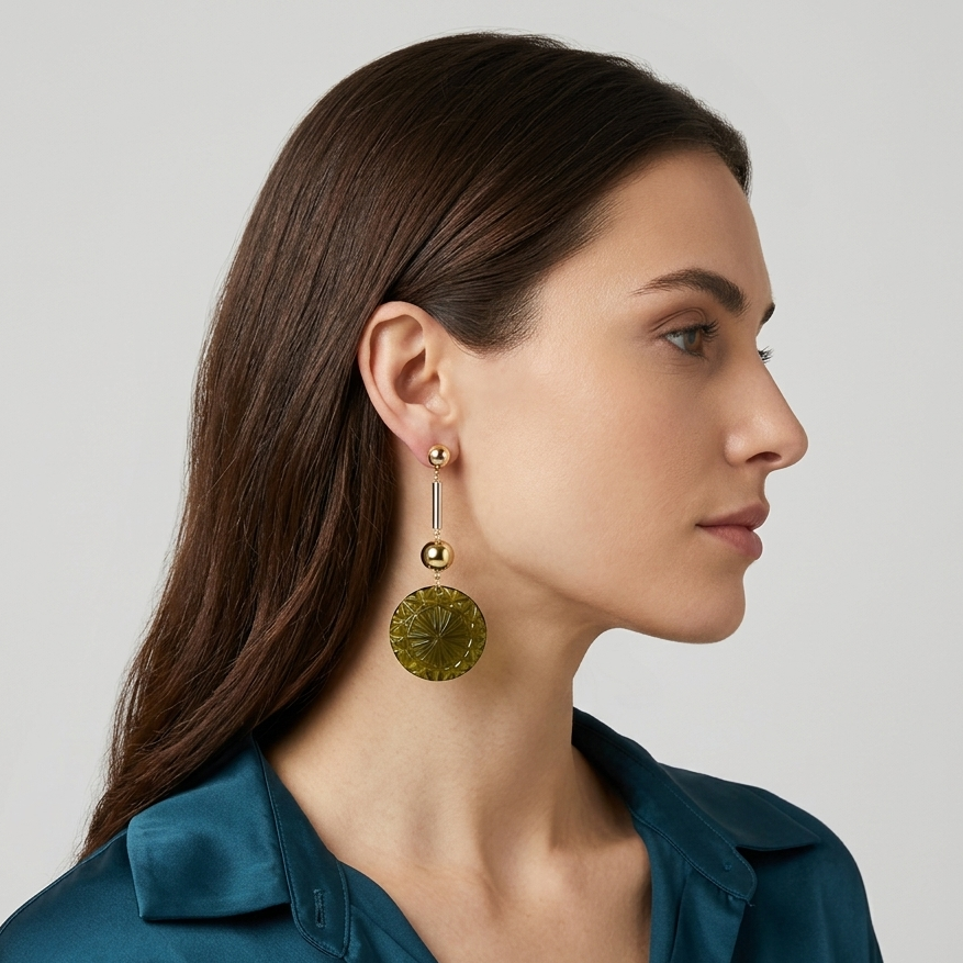 A woman in profile wearing a pair of gold drop earrings with a small bead and a larger textured circular disc, paired with a teal satin blouse against a light gray background.