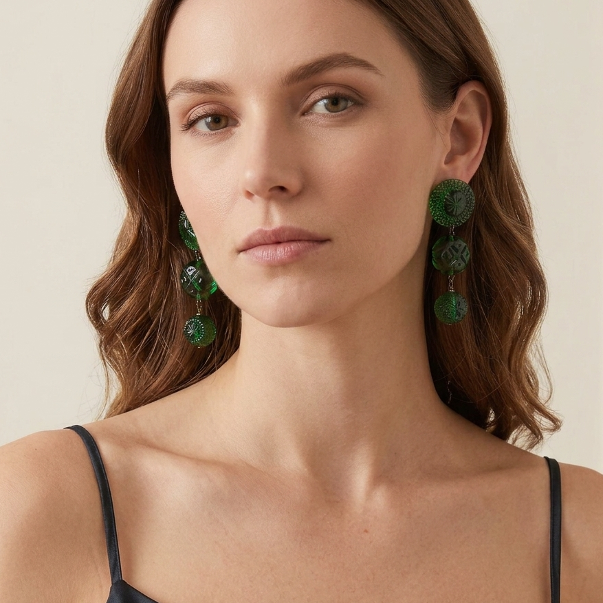 A woman wearing large green circular drop earrings with layered circular details, paired with a black spaghetti-strap top against a soft beige background.