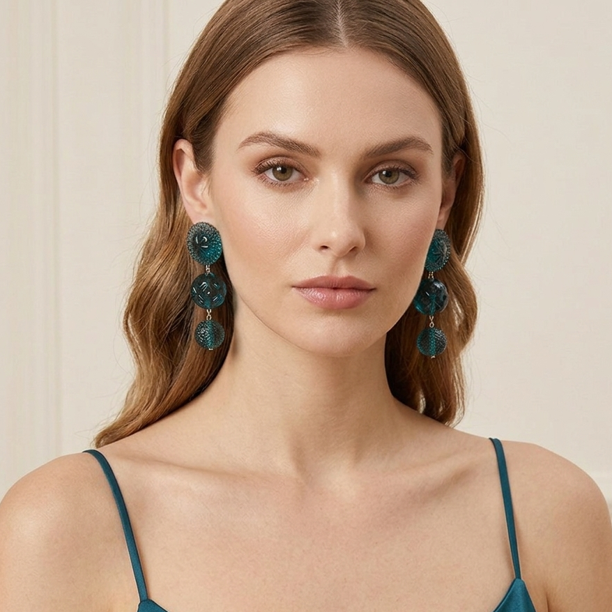 A woman wearing large, multi-tiered teal drop earrings with a glossy finish, paired with a matching teal camisole top against a soft cream-colored background.