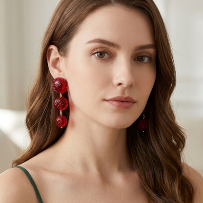 A woman with long wavy brown hair wearing a dark green spaghetti-strap top and matching red drop earrings, photographed in a softly lit indoor setting.