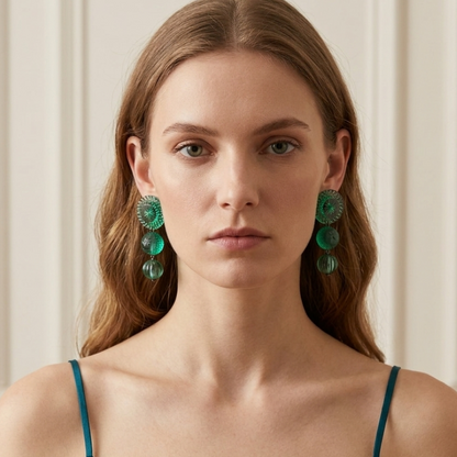 A woman with light brown hair wearing large, chunky green beaded drop earrings and a teal spaghetti-strap top, set against a soft beige paneled wall.