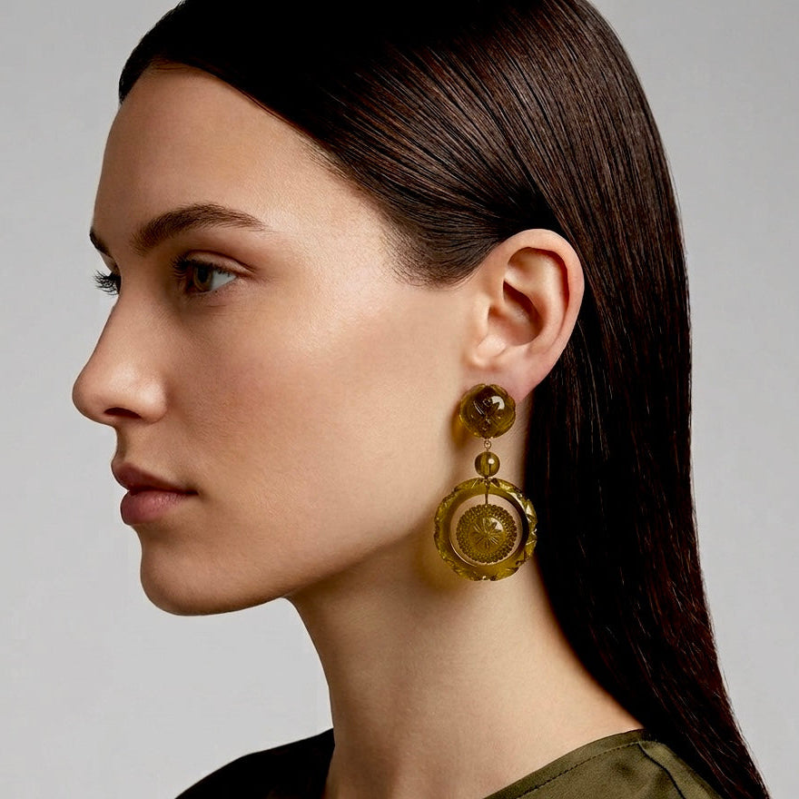 A close-up side profile of a woman wearing a large, circular gold-toned dangle earring with a textured, amber-gold center, set against a neutral gray background.