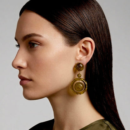 A close-up side profile of a woman wearing a large, circular gold-toned dangle earring with a textured, amber-gold center, set against a neutral gray background.
