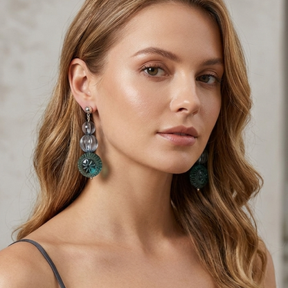 A woman with wavy light brown hair wearing a dark gray spaghetti-strap top and long, dangling teal and silver statement earrings, photographed in a softly lit indoor setting.
