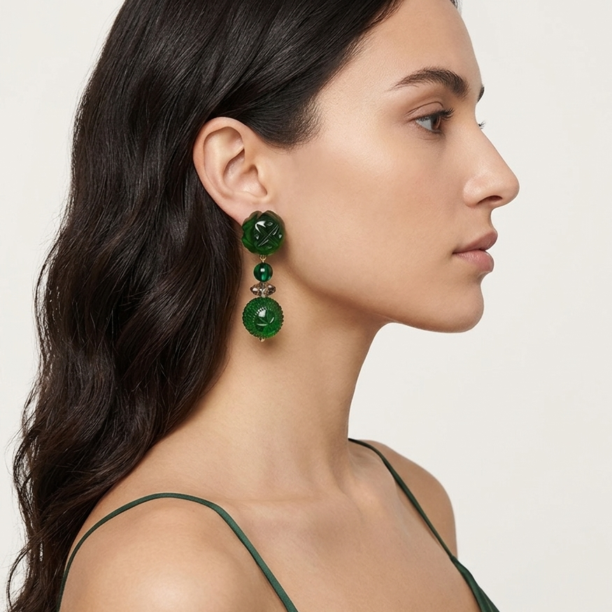 A woman in profile wearing a pair of green drop earrings with layered circular and oval beads, paired with a dark green satin top against a light off-white background.