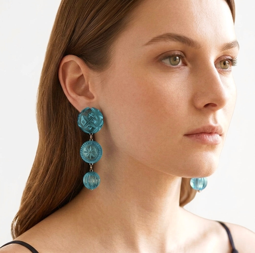 A close-up of a woman wearing a pair of dangling earrings made of three stacked, round turquoise-blue translucent beads with a textured center, set against a clean white background.