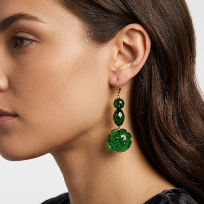 A close-up side profile of a woman wearing a single drop earring made of three stacked green faceted beads with a larger flower-shaped bead at the bottom, set against a plain light gray background.