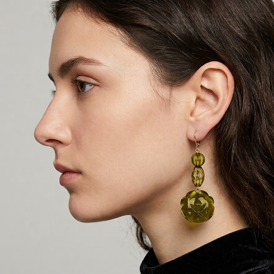A close-up side profile of a woman wearing a long, faceted olive-green dangle earring with two stacked beads and a larger round pendant, paired with a black velvet top against a neutral gray background.