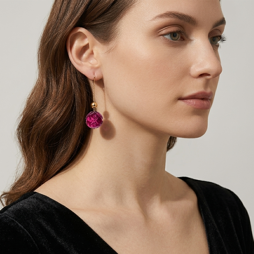 A close-up of a woman wearing a dangling earring with a faceted magenta-pink gemstone and a gold-toned post, paired with a black velvet top against a light gray background.