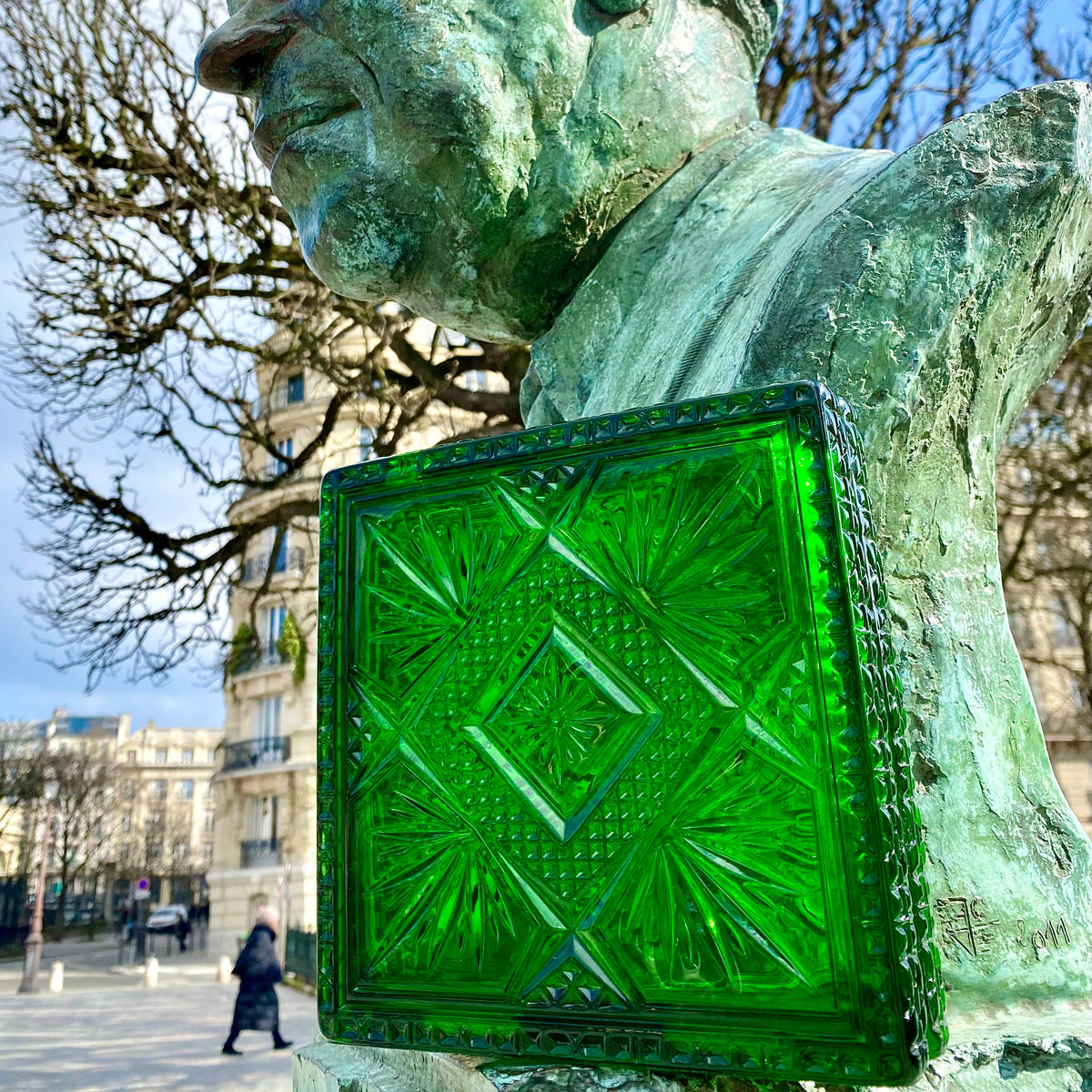 A close-up outdoor shot of a green patina bronze bust sculpture with a large, decorative green glass panel attached to its side, set against a city square background with trees and a blue sky.