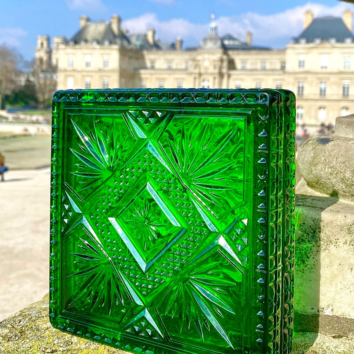 A square green cut-glass inspired clutch with a geometric diamond pattern, standing on a stone ledge with a blurred classical building and blue sky in the background.