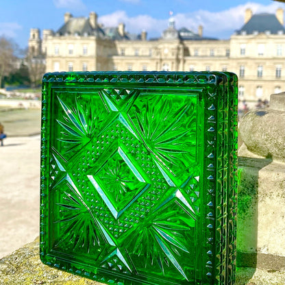 A square green cut-glass inspired clutch with a geometric diamond pattern, standing on a stone ledge with a blurred classical building and blue sky in the background.