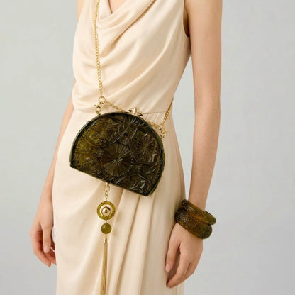 A model wearing a cream-colored draped dress carries a small, dark olive-green embossed handbag with a gold chain strap and tassel.