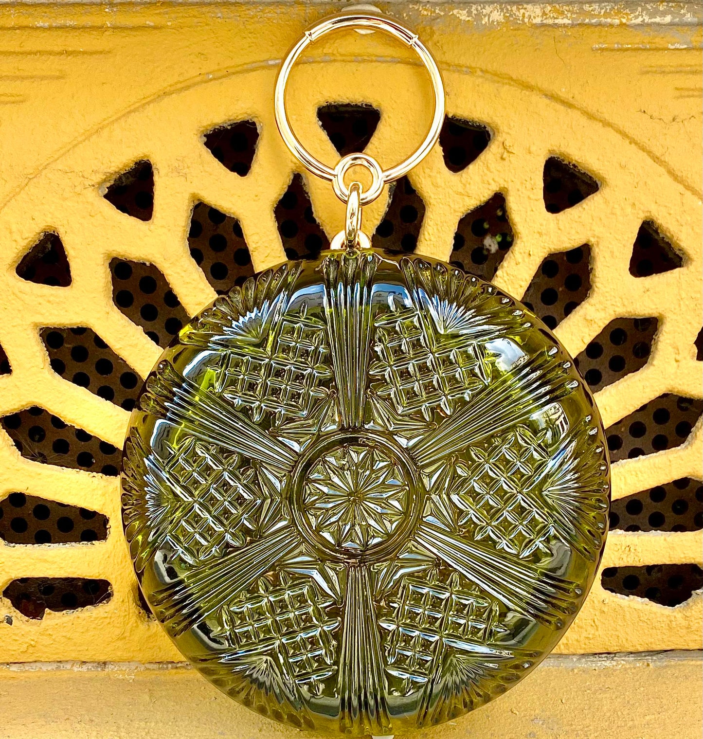 A round, textured olive-green clutch bag with hand carved intricated patterns with a gold tone ring handle, displayed against a yellow decorative background.