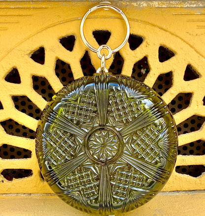 A round, textured olive-green clutch bag with hand carved intricated patterns with a gold tone ring handle, displayed against a yellow decorative background.